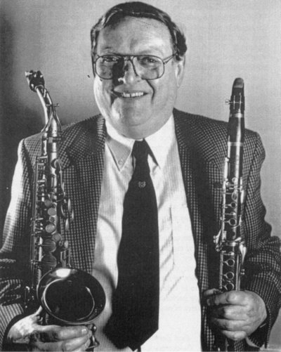 An older man with glasses smiling, holding a saxophone in one hand and a clarinet in the other