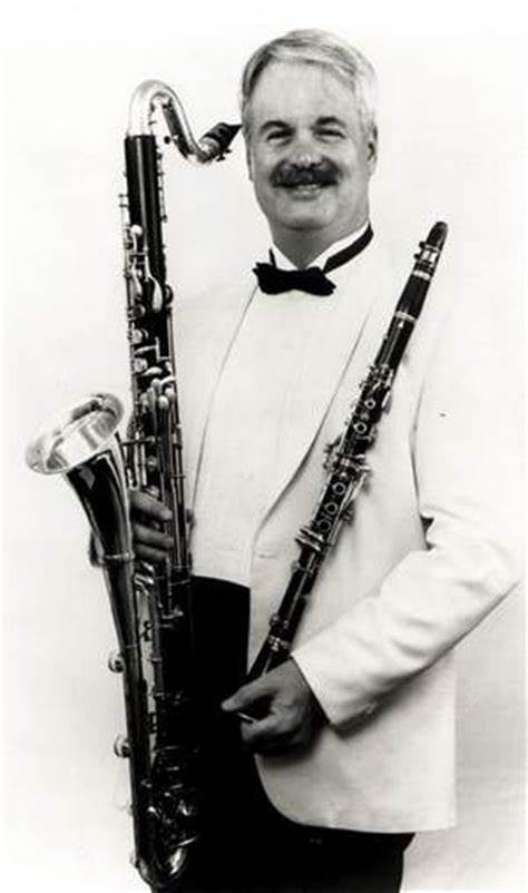 An older man with a moustache smiling, holding a bass clarinet in one hand and a Bb clarinet in the other