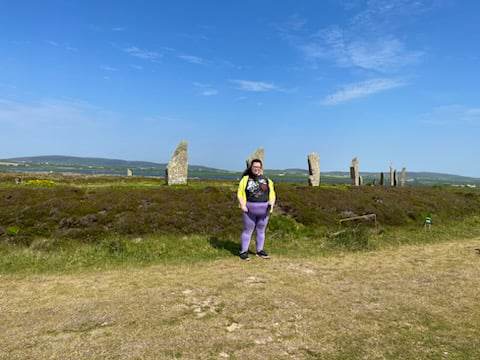 A picture of me wearing a tshirt with Thor on it and a yellow hoodie, smiling in the sun. I am standing in front of the Ring of Brodgar, a large stone circle in Orkney