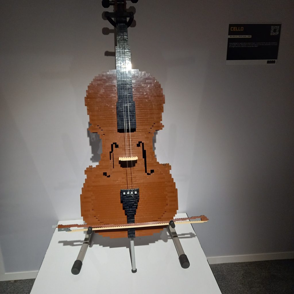 A large brown cello sits on a white stand with a bow at the bottom, all made of lego 